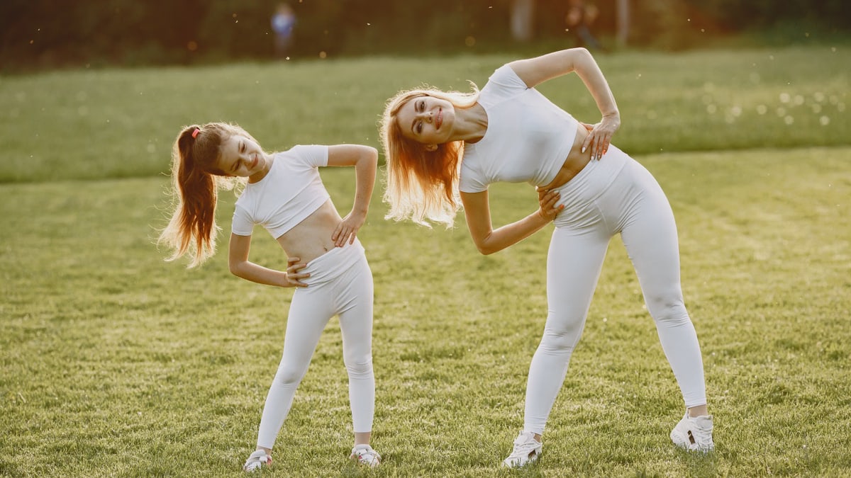 A mother and daughter in activewear stretching on a sunny day in a grassy field.